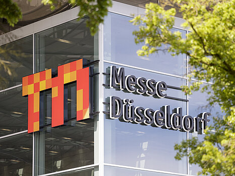 "Messe Düsseldorf" logosu, yeşil ağaç dallarıyla kısmen çerçevelenmiş olarak cephede göze çarpan, modern bir cam binanın dış görünümü.